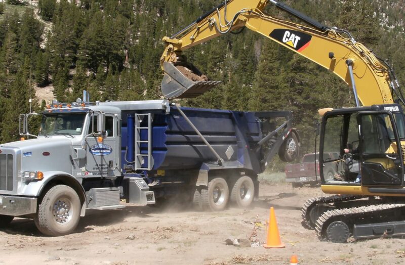 excavator lifting dirt into dumptruck by forest