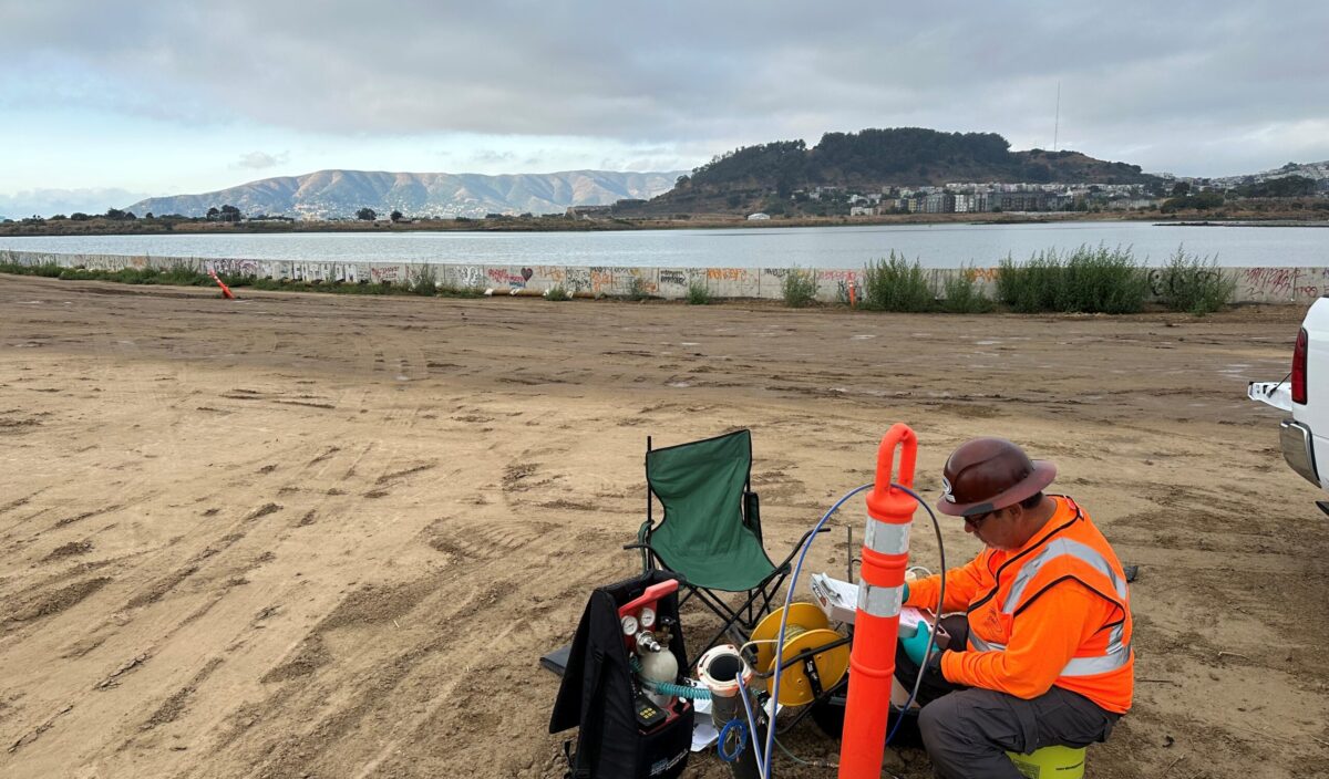 HP_ParcelE2Sampling person working with equipment in sandy area near water and mountains in horizon